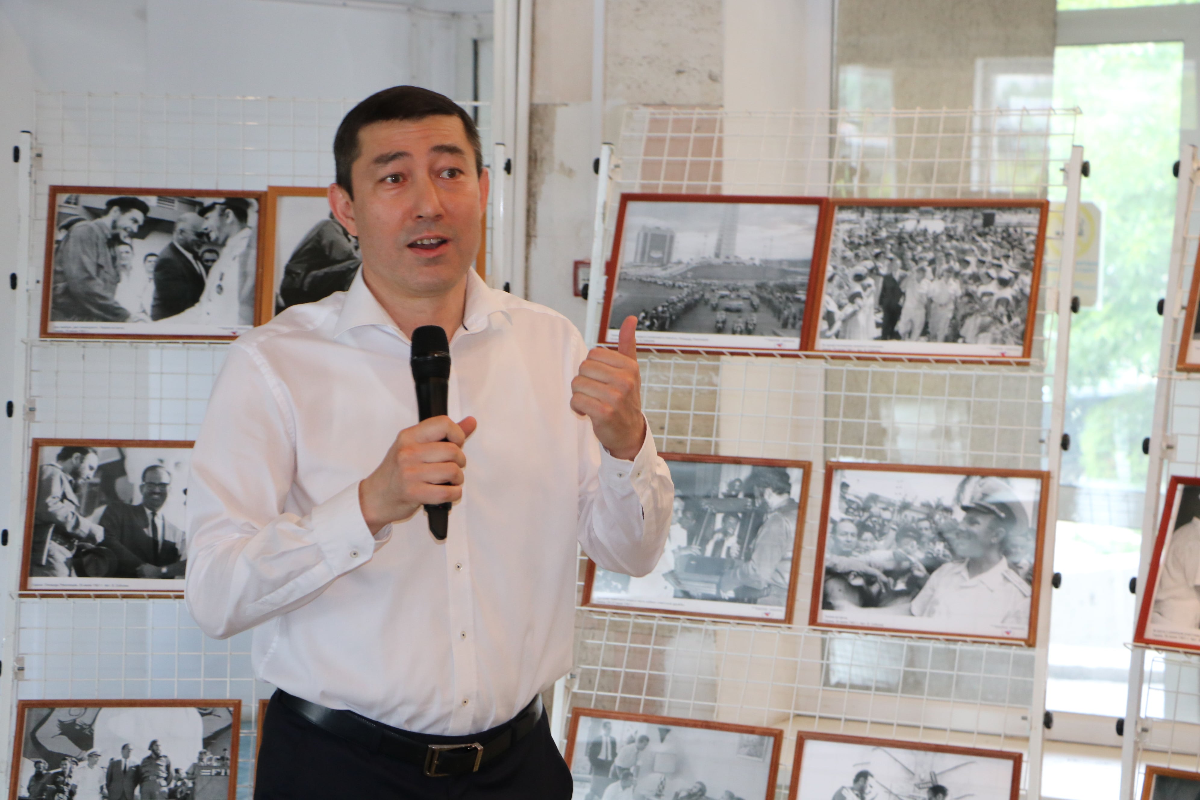 На фото: человек в белой рубашке выступает с микрофоном на фоне стэнда с фотографиями.