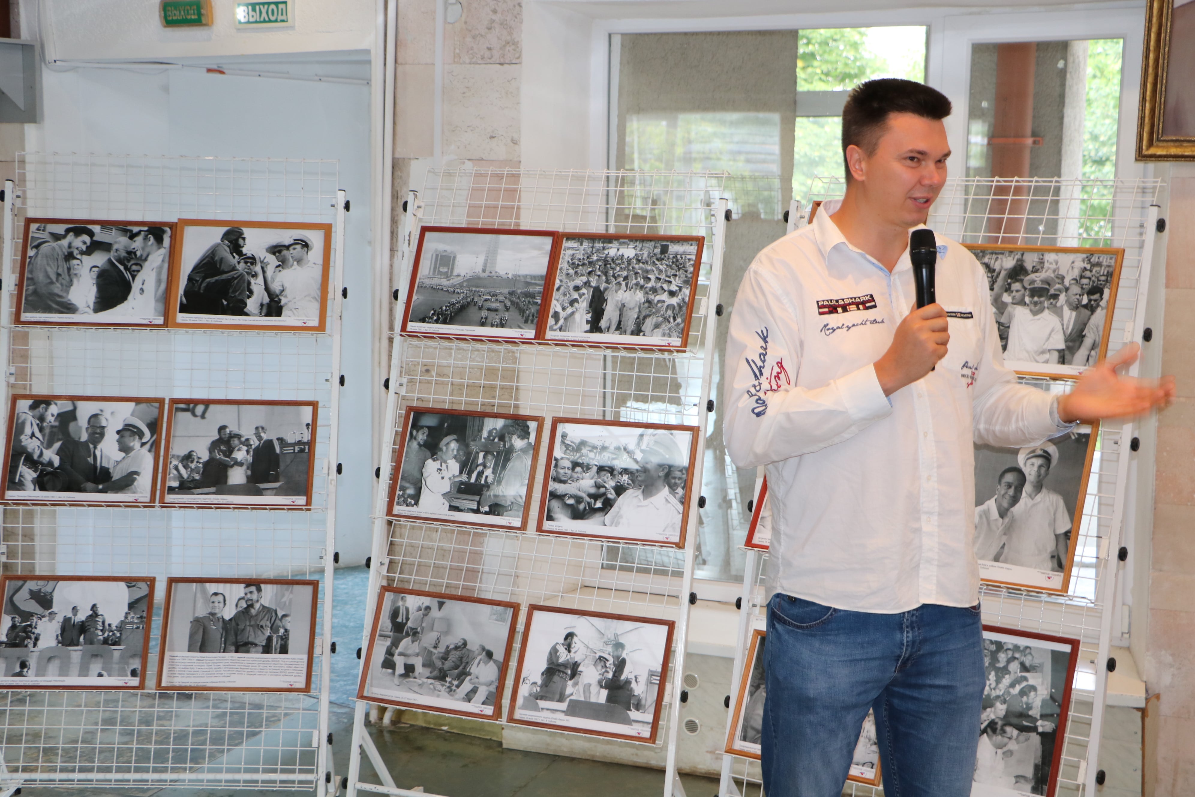 На фото: Алексей Андруцкий в белой рубашке выступает с микрофоном на фоне стэнда с фотографиями.